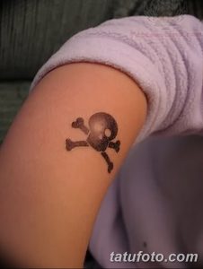 Фото татуировки на плече с изображением черепа и костей, выполненной в коричневых тонах. Небольшой и лаконичный рисунок.