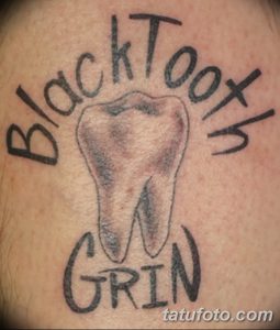 Фото тату зуба на спине с надписью 'BlackTooth Grin'. Черно-серая татуировка в стиле леттеринг, с акцентом на текст и детализацию зуба.