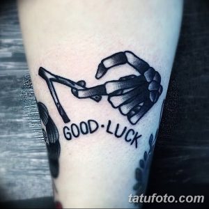 Фото тату: черно-белая татуировка рукопожатия двух скелетов с надписью "GOOD LUCK" на предплечье. Изображение выполнено в минималистичном стиле.
