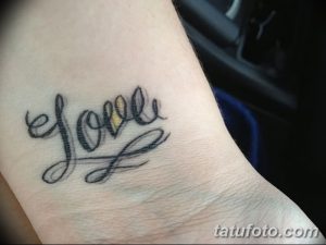 Фото тату на запястье: надпись 'Love' с сердечком вместо буквы 'o'. Черно-серый рисунок татуировки, выполненный каллиграфическим шрифтом.