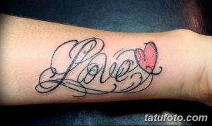 Фото тату надписи 'Love' с красным сердцем и декоративными элементами на предплечье. Рисунок татуировки в стиле каллиграфии.