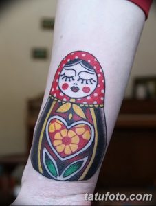 Фото тату матрешки с закрытыми глазами, украшенной сердечком и цветами. Милый рисунок татуировки для девушек, любящих нежные мотивы.