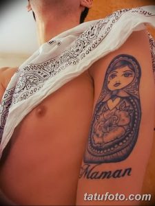 Фото тату матрешки на груди мужчины. Рисунок выполнен в черно-серых тонах с добавлением красных акцентов. Под матрешкой надпись 'Maman'.