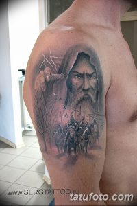 Фото тату: реалистичное изображение воина с бородой и грозным взглядом, окруженного молниями и деревьями на плече. Детализированный рисунок в черно-серых тонах.