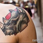 Фото тату носорога в стиле олд скул на плече. Рисунок выполнен в ярких цветах с использованием четких контуров и красных акцентов.