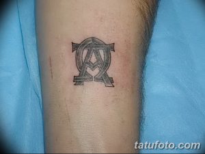 Черно-серое фото татуировки с буквами Альфа и Омега, расположенными в стилизованном кресте на предплечье. Классический и религиозный дизайн.