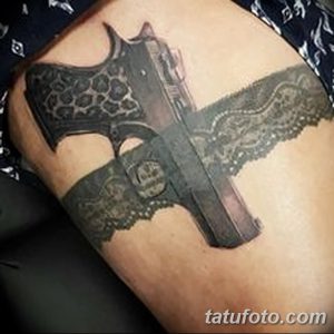 Фото тату кружевной повязки с пистолетом на ноге. Детализированный рисунок татуировки в стиле реализм, выполненный в черно-серых тонах.