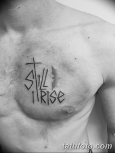 Фото тату: надпись "Still I Rise" с изображением креста и крыльев, выполненная на груди. Татуировка символизирует силу духа и преодоление трудностей.
