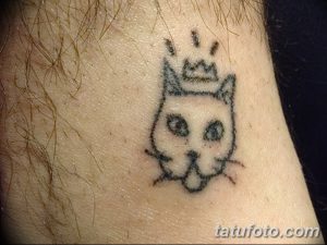 Фото тату на лодыжке: небольшая, слегка размытая татуировка кошачьей мордочки с короной. Рисунок татуировки выполнен в серо-черных тонах.