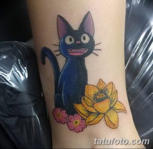 Фото тату на руке с изображением черного кота Джиджи из аниме Kiki's Delivery Service. Рисунок татуировки выполнен в ярких цветах с использованием желтых нарциссов.