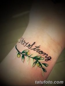 Фотография татуировки на запястье с надписью "Stay Strong" и изображением ветки с зелеными листьями. Черно-зеленый рисунок в стиле каллиграфия.