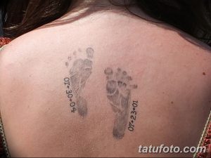 Фото тату с двумя отпечатками детских ножек и датами рождения на спине. Черно-серый рисунок татуировки, выполненный в реалистичном стиле.
