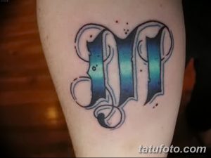 Фото тату: стилизованная буква 'M' в ярких голубых и черных тонах, выполненная в стиле олд скул на предплечье.