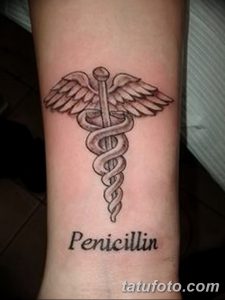 Фото тату Кадуцея с крыльями и надписью 'Penicillin' на предплечье. Черно-белый рисунок татуировки, выполненный в классическом стиле.