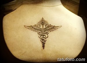 Фото тату: Небольшая коричневая татуировка с изображением посоха Эскулапа на лопатке. Элегантный рисунок татуировки для женщин.