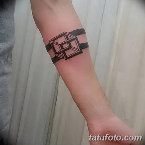 Фотография минималистичной татуировки на предплечье, изображающей черный куб, расположенный над черной полосой, создающей эффект браслета.