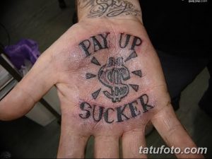 Фото татуировки на ладони с надписью "PAY UP SUCKER" и изображением доллара с крыльями. Рисунок выполнен в черном цвете, с использованием четких линий и контрастных цветов.