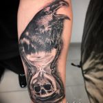 Фото тату песочные часы на предплечье, украшенные вороном и черепом. Детализированный черно-серый рисунок татуировки в реалистичном стиле.