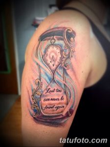 Фото тату песочные часы на плече, украшенные циферблатом и надписью 'Lost time can never be found again'. Цветной рисунок татуировки в реалистичном стиле.
