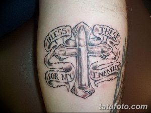 Фото тату: стилизованный крест с лентами и надписью 'Bless These For My Enemies', выполненная в черно-белом стиле на руке.