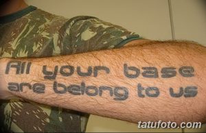 Фото тату на предплечье: фраза 'All your base are belong to us', выполненная простым шрифтом в сером цвете.