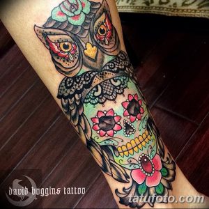 Фото тату совы, стилизованной под сахарную черепушку (calavera), с яркими цветами и узорами на ноге. Использованы цвета: розовый, бирюзовый, черный.