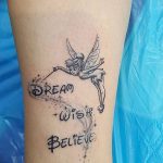 Фото тату феи Tinkerbell, держащей волшебную палочку, с надписью "Dream, Wish, Believe" на руке, выполненное в черно-белой гамме.