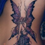 Фото тату: мистическая фея с большими крыльями, сидящая среди грибов и ветвей. Детализированный рисунок татуировки в темных фиолетовых тонах.