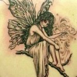 Черно-серая татуировка феи, сидящей на ветке дерева и держащей в руках цветок. Детализированный рисунок с акцентом на крылья и лицо.