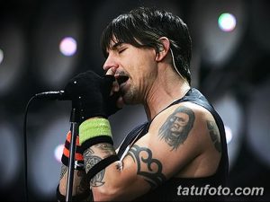 Фотография музыканта, выступающего на сцене с татуировками на руках и плечах. Мужчина с микрофоном и страстным выражением лица.