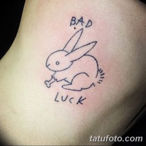 Фото тату кролика с костью и надписями 'BAD' и 'LUCK' на руке. Милый минималистичный рисунок татуировки в черном цвете.