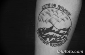 Фото татуировки на ноге мужчины: изображение гор в круге с надписью "Достигнув вершины. Продолжай восхождение". Черно-белый рисунок в стиле графика.
