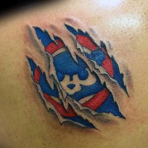 Фото тату на плече с изображением разорванной эмблемы Chicago Cubs, сквозь которую видна кожа. Реалистичный рисунок с использованием синих и красных цветов.