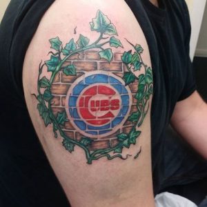 Фото тату на плече с логотипом Chicago Cubs на фоне кирпичной стены, увитой плющом. Яркие цвета и детализированный рисунок.