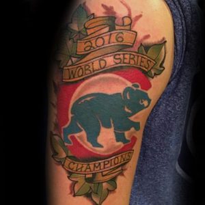Фото тату на плече с изображением медведя, символа Chicago Cubs, на фоне красного баннера с надписью 'World Series Champions 2016'.