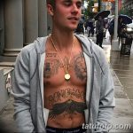 Фотография Джастина Бибера в толстовке, демонстрирующая его татуировки на груди и животе. Видны крест, надпись 'SONOFGOD' и другие рисунки.