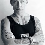 Фото тату на теле мужчины: рукава с рисунками, грудь с татуировками. Мужчина стоит, скрестив руки.