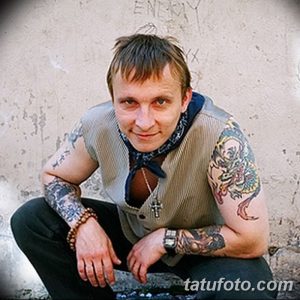 Фотография Игоря Талькова, сидящего на фоне стены, с татуировками на руках и груди.