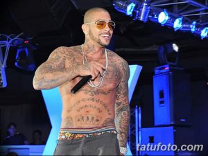 Фотография мужчины с бритой головой и татуировками на теле, выступающего на сцене с микрофоном. Он одет в черную майку и золотую цепь.