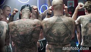 Фотография двух мужчин с бритыми головами и татуировками на теле, стоящих в клубе. Они демонстрируют татуировки на спине.