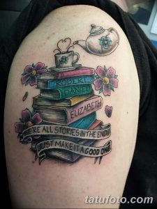 Фото татуировки на плече с изображением стопки книг, украшенной цветами и цитатой, выполненной в ярком стиле.