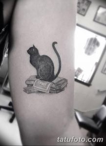 Фото тату на предплечье: черный кот, сидящий на стопке книг и читающий книгу. Черно-белый рисунок с детальной прорисовкой.