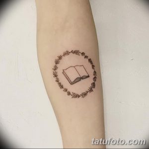 Фото тату: небольшая татуировка с изображением открытой книги в окружении венка из листьев. Черно-белый рисунок, минималистичный стиль.