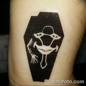 Фото татуировки на руке с изображением гроба, внутри которого находится сюрреалистичное лицо с глазами и скелетом. Черно-белый рисунок в абстрактном стиле.