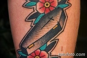 Яркая татуировка на руке с изображением гроба, украшенного цветами. Выполнена в традиционном стиле олдскул с использованием четких контуров.