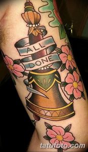 Фото татуировки колокола, украшенного цветами и лентой с надписью "ALL DONE" на руке. Красочный и детализированный рисунок татуировки в стиле олдскул.