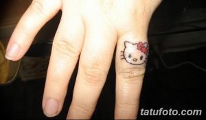 Фото тату: крошечная татуировка Hello Kitty на пальце руки. Изображение выполнено в черном цвете с красным бантиком.
