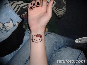 Фото тату: маленькая татуировка Hello Kitty на запястье. Изображение выполнено в черном цвете с красным бантиком.