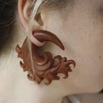 Фото деревянного плага для уха с резным узором. Плаг коричневого цвета, украшен сложным растительным орнаментом, вставлен в растянутое ухо.