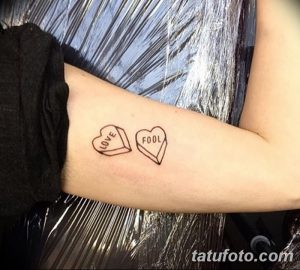 Фото татуировки с двумя сердцами, на одном из которых написано 'Love', а на другом 'Fool'. Черно-белый рисунок татуировки на предплечье.